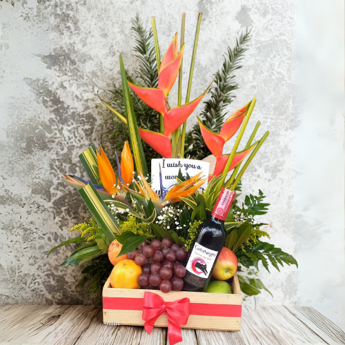 Arreglo floral tropical con heliconias y flores naranjas acompañado de uvas, peras, manzanas y una botella de vino tinto en caja de madera con lazo rojo