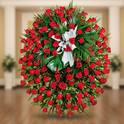 Arreglo floral funerario en forma de corona ovalada con numerosas rosas rojas y hojas verdes, decorado con lazo blanco sobre fondo interior desenfocado
