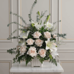 Arreglo de rosas color crema y lirios blancos con follaje verde en maceta blanca sobre mesa de mármol