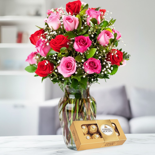Ramo de rosas rojas y rosas en jarrón de cristal junto a caja de bombones dorada sobre mesa blanca