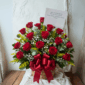 Canasta de mimbre con arreglo de rosas rojas, flores blancas pequeñas y follaje verde, decorada con lazo rojo y tarjeta sobre fondo claro