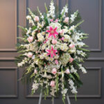 Arreglo floral de pie con lirios rosas al centro, rosas rosadas y flores blancas rodeadas de follaje verde sobre fondo de pared gris