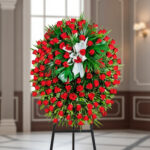 Arreglo floral en forma de corona ovalada con numerosas rosas rojas y follaje verde sobre caballete negro en interior elegante