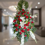 Arreglo floral fúnebre en forma ovalada con rosas rojas, lirios blancos y follaje verde sobre fondo de interior elegante desenfocado