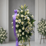 Arreglo floral funerario en forma de corona con rosas y lirios blancos, decorado con cinta morada, sobre caballete en interior elegante