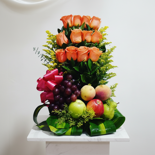 Arreglo vertical de rosas naranjas combinado con frutas frescas como uvas moradas, manzanas verdes y rojas y duraznos, decorado con hojas verdes y un lazo rosa sobre pedestal blanco