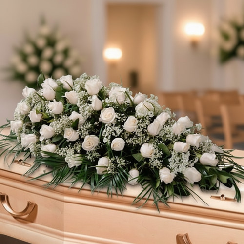 Centro de flores funerarias con rosas blancas y follaje verde colocado sobre un ataúd de madera en una sala velatoria iluminada