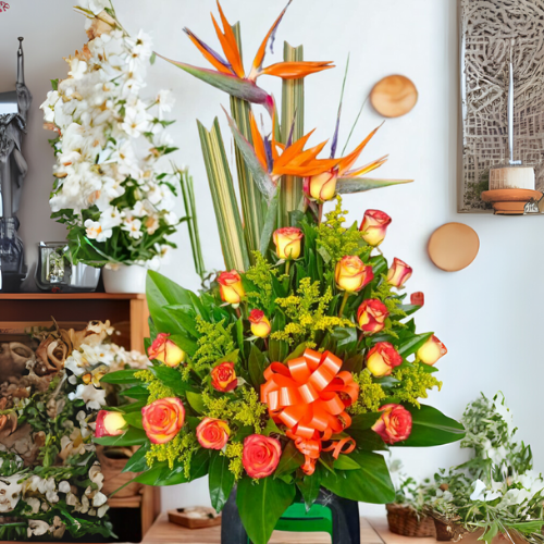Arreglo floral exótico con aves del paraíso naranjas y rosas bicolor amarillas y naranjas, decorado con lazo naranja sobre base verde en interior de floristería