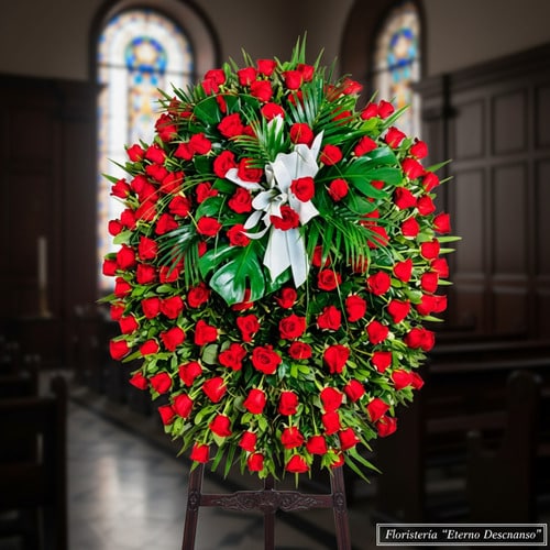 Arreglo floral funerario ovalado con numerosas rosas rojas y follaje verde, lazo blanco al centro, colocado sobre caballete de madera dentro de una iglesia