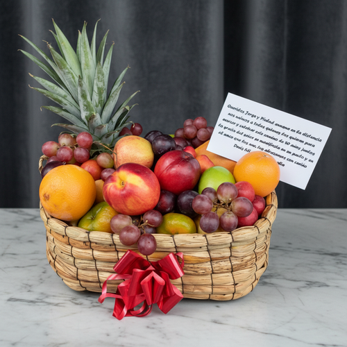 Canasta de frutas variadas con tarjeta sobre mesa de mármol Canasta tejida llena de frutas frescas como piña, naranjas, manzanas, nectarinas, uvas y limones, decorada con lazo rojo y tarjeta de dedicatoria sobre mesa de mármol con fondo gris