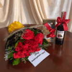 Arreglo de regalo con ramo de rosas rojas, botella de vino tinto con lazo rojo, caja de chocolates y tarjeta de felicitación sobre mesa de madera