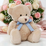 Osito de peluche color beige con lazo de cuadros celeste y blanco sentado frente a un ramo de rosas rosadas y blancas desenfocado