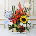 Arreglo de flores tropicales y girasol combinado con frutas frescas en caja blanca y globo que dice Feliz Día