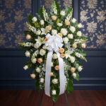 Arreglo funerario en forma de corona con rosas blancas y crema, lirios blancos y follaje verde, decorado con gran lazo blanco sobre caballete metálico