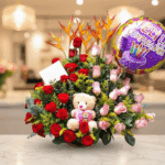 Arreglo de flores con rosas rojas y rosas, flores exóticas naranjas, peluche de oso y globo de feliz cumpleaños sobre mesa en interior