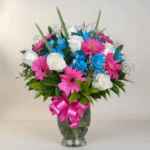 Ramo de flores en jarrón de cristal con rosas blancas, gerberas rosas, flores azules y lazo rosa brillante
