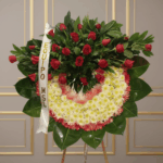 Arreglo fúnebre en forma de corona con rosas rojas en la parte superior y flores blancas y rosas en la parte inferior, sobre base de hojas verdes y cinta con texto Equipo MSL