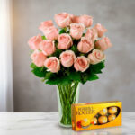 Arreglo de rosas color rosa en florero de vidrio transparente acompañado de una caja de chocolates Ferrero Rocher sobre superficie blanca