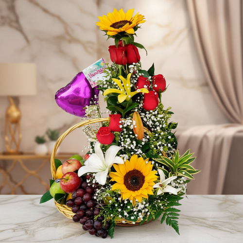 Arreglo floral con girasoles, rosas y frutas en canasta Canasta de mimbre con arreglo de girasoles, rosas rojas, lirios blancos y amarillos, globo morado en forma de corazón y frutas como uvas y manzanas sobre mesa de mármol