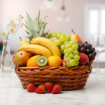 Cesta de mimbre llena de frutas frescas como plátanos, uvas, piña, naranjas, fresas y kiwi sobre una mesa de mármol en una cocina luminosa