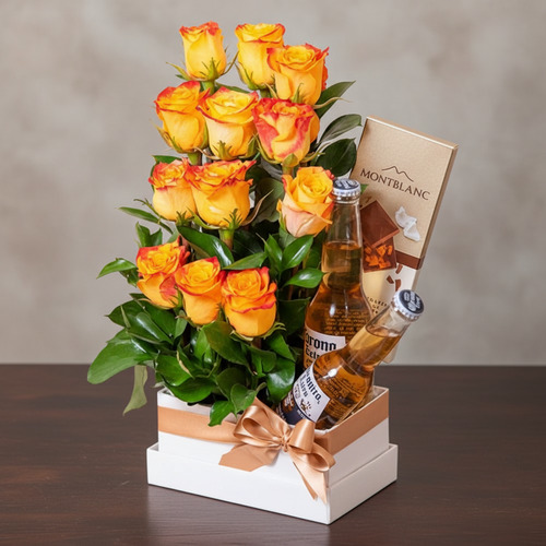 Arreglo de rosas amarillas y naranjas en caja blanca con lazo beige, acompañado de dos botellas de cerveza Corona y una barra de chocolate Montblanc sobre mesa de madera