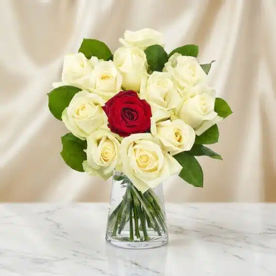 Arreglo floral CARLA con 12 rosas de tallo largo, una roja central y once blancas, en un jarrón de cristal sobre mármol.