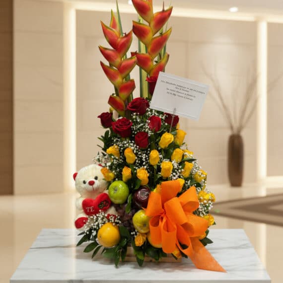 Arreglo Floral con Frutas Encanto: Elegancia y Frescura para Regalar Arreglo floral premium con rosas rojas y amarillas, heliconias, frutas frescas y un lazo naranja, sobre una mesa de mármol en
