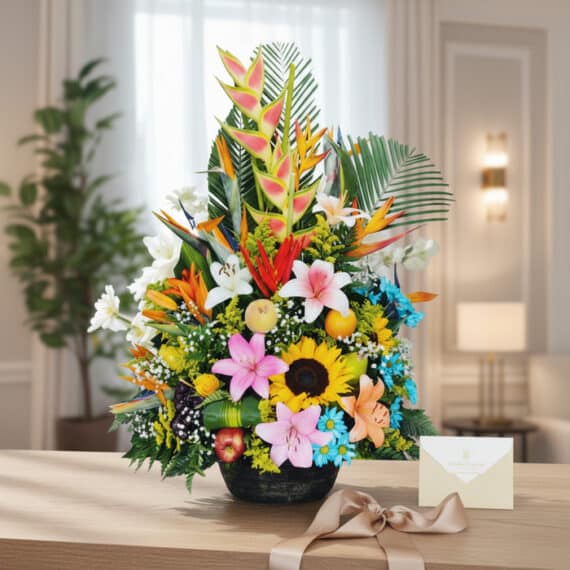 Arreglo floral exótico con heliconias, lirios, girasoles y frutas frescas, presentado sobre una mesa de madera con una tarjet
