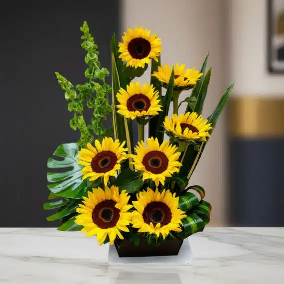 Arreglo floral premium con girasoles Bengala, hojas verdes y follaje, sobre una mesa de mármol en un lobby de hotel boutique.