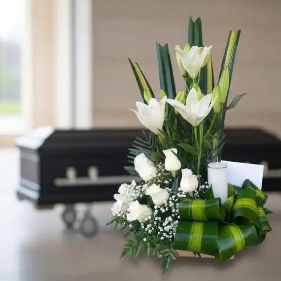 Arreglo floral fúnebre Claridad con lirios y rosas blancas, vela y tarjeta, en sala de velación moderna con ataúd al fondo.