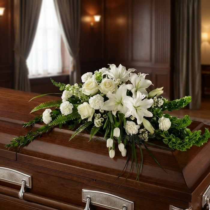 Arreglo floral fúnebre cubre caja 'Bondad' con lirios, rosas y claveles blancos, sobre un ataúd de madera oscura en una sala