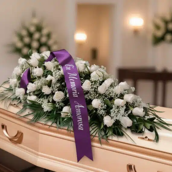 Arreglo fúnebre cubre caja Cielo con rosas blancas y follaje verde, adornado con una cinta morada que dice 'En Memoria Amoros