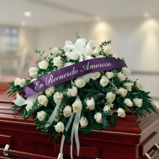 Arreglo fúnebre cubre caja Misericordia con rosas blancas y follaje verde, adornado con una cinta morada que dice 'En Recuerd
