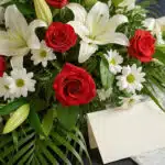 Arreglo fúnebre cubre caja Paraíso con rosas rojas, lirios blancos y margaritas, con rocío sutil y follaje verde, vista macro