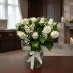 Arreglo fúnebre Pétalos del Alma con rosas blancas y follaje verde en un jarrón de cristal con lazo blanco, sobre una mesa de