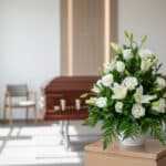 Arreglo floral fúnebre Piedad con rosas y lirios blancos en sala de velación moderna, ataúd desenfocado al fondo.