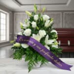 Arreglo fúnebre Piedad con rosas y lirios blancos, cinta morada con mensaje de condolencia, en sala de velación premium.