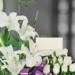 Primer plano de Arreglo Fúnebre Pureza con lirios blancos, rosas y cinta morada, rocío sutil en pétalos y follaje, profundida