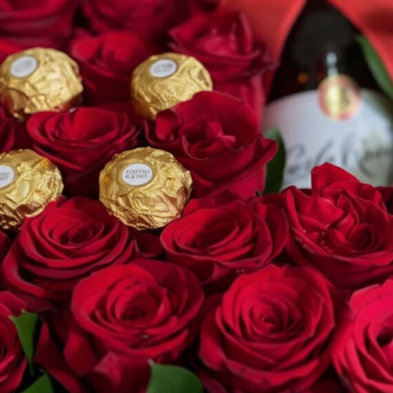Primer plano extremo de la Caja Floral ENRICO con rosas rojas frescas, chocolates Ferrero Rocher y una botella de vino, desta