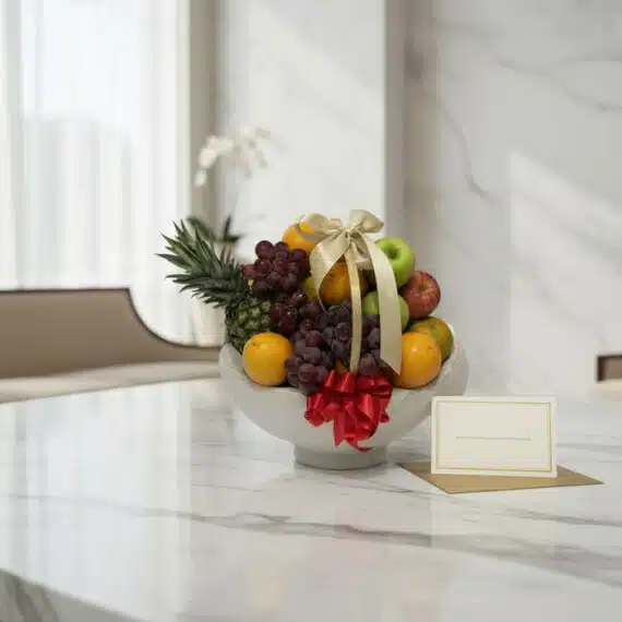 Canasta de frutas gourmet con piña, uvas, manzanas y naranjas, decorada con cintas doradas y rojas, sobre una mesa de mármol