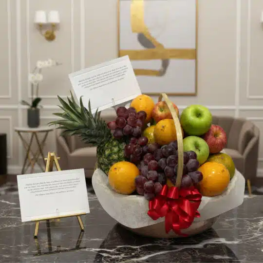 Canasta de frutas premium con piña, uvas, manzanas rojas y verdes, naranjas, y un lazo rojo, sobre una mesa de mármol en un e
