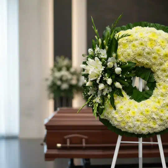 Corona fúnebre Bethesda de crisantemos amarillos y flores blancas, en sala de velación moderna con ataúd desenfocado al fondo