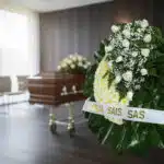 Corona fúnebre Descansa en Paz con rosas blancas y follaje verde, cinta personalizada, en sala de velación moderna con ataúd