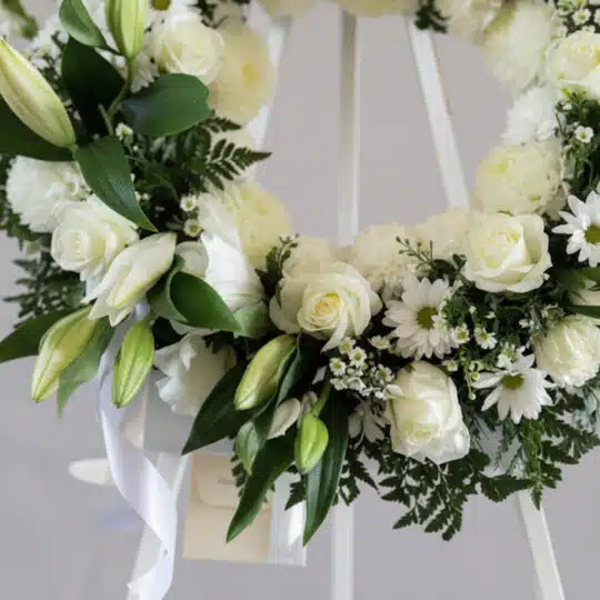 Corona fúnebre 'Homenaje Sincero' con lirios, rosas y crisantemos blancos, follaje verde, rocío sutil, cinta satinada y sobre