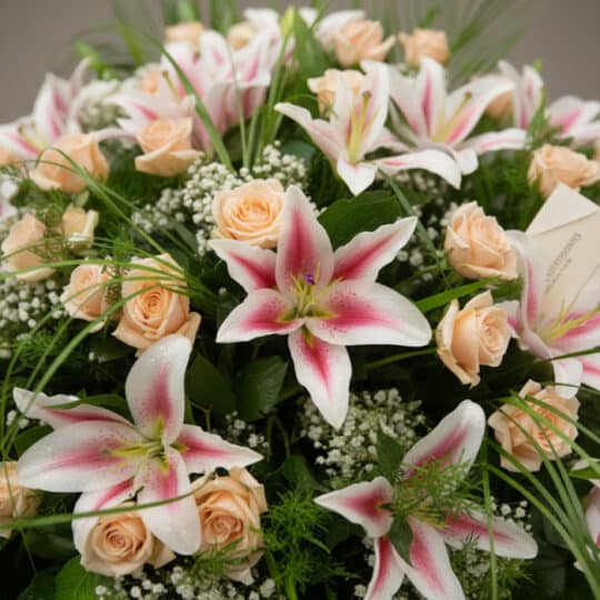 Primer plano de una corona fúnebre 'Luz Eterna' con lirios blancos y rosas melocotón, rocío sutil, cinta satinada y follaje v