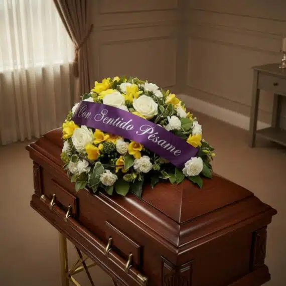 Cubre caja fúnebre Baltasar con rosas blancas, fresias amarillas y follaje verde, adornado con una cinta morada que dice 'Con