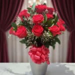 Jarrón plateado con doce rosas rojas frescas y follaje verde, atado con un lazo rojo brillante, sobre una mesa de mármol blan