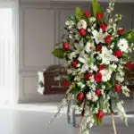 Pedestal fúnebre Adoración con lirios blancos, rosas rojas y gerberas blancas en sala de velación moderna, transmitiendo resp