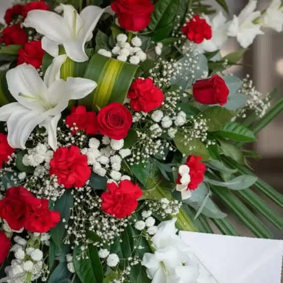 Primer plano de un pedestal fúnebre Homenaje con rosas rojas, lirios blancos y claveles, con rocío sutil y cinta satinada.